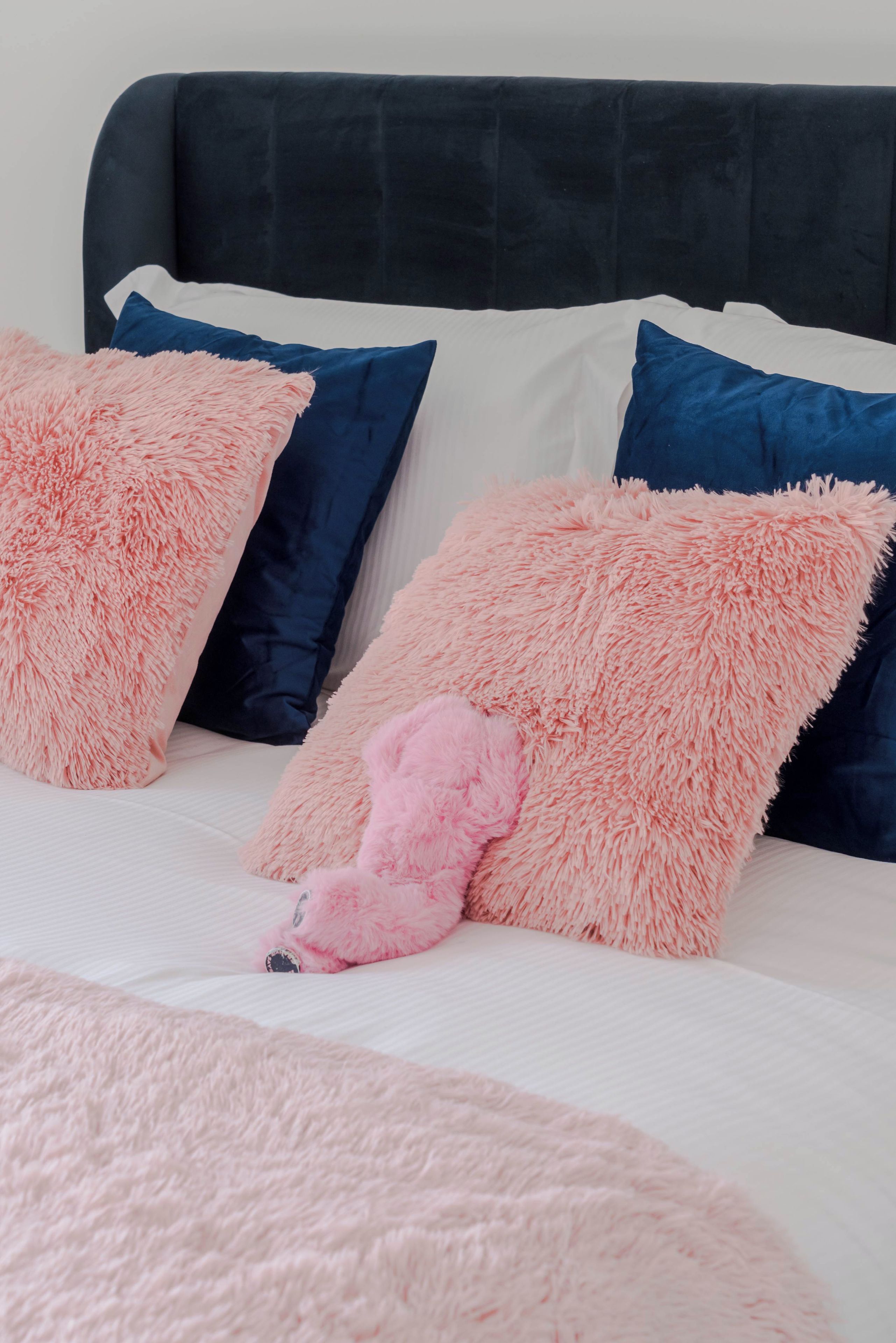 A bed with a dark headboard, white bedding, and fluffy pink and blue pillows.