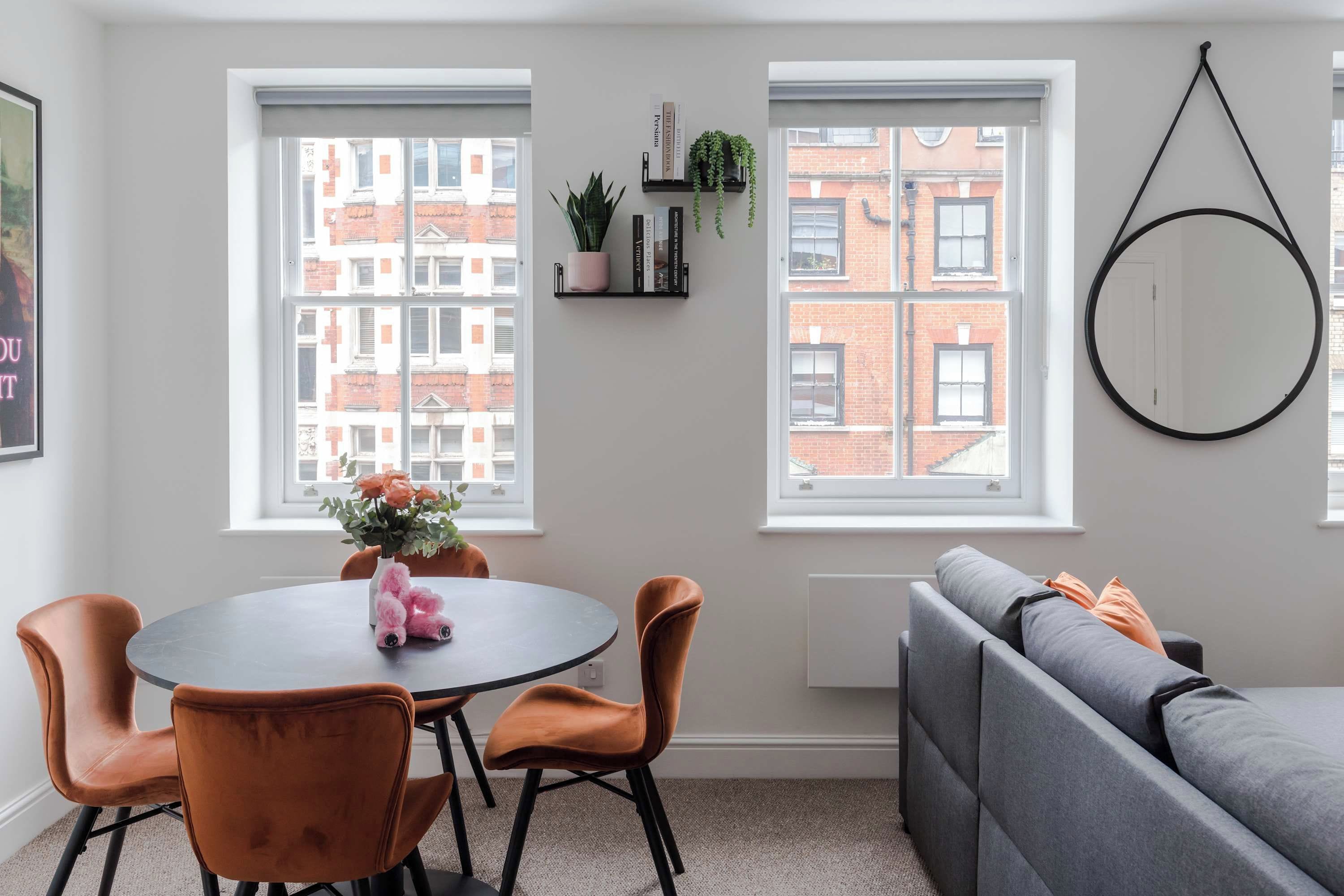 A dining area with a round table, four chairs, a sofa, and two windows with a view of buildings.