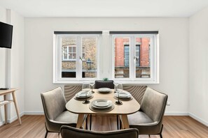 A dining area with a table set for four, two windows with brick buildings outside, a flat-screen TV on the wall, and a wooden floor. - Covent Garden Studio · Sleeps 3 · Floral St (London)