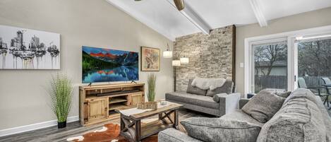 A living room with a ceiling fan, a flat-screen TV, a stone wall, a gray sofa, and a cowhide rug.