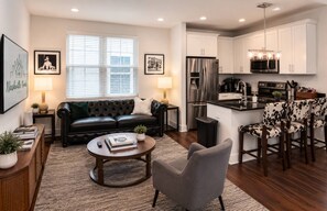 A modern living room with a black leather sofa, a grey armchair, and a coffee table. There is a kitchen area with white cabinets and stainless steel appliances. - Sleeps 10 Near Broadway | Hart Suites Unit #2 (Nashville)