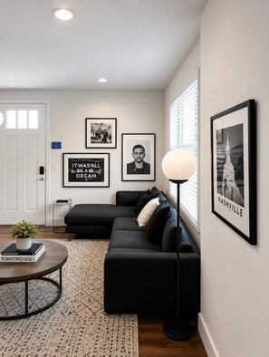 Ein modernes Wohnzimmer mit einer Eckcouch, einem runden Couchtisch und einer Stehlampe. An der Wand befinden sich gerahmte Bilder, eine Tür mit Fenster und ein gemustertes Teppich.