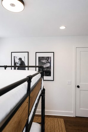 Ein Etagenbett mit Holz-Kopfteil, eine weiße Wand mit zwei gerahmten Schwarz-Weiß-Fotos, eine weiße Tür und ein brauner Teppich aus Gewebe auf dem Boden.