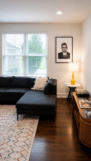 Ein Wohnzimmer mit einem dunkelgrauen Sofa, einem gemusterten Teppich, einem kleinen Tisch mit einer gelben Lampe und einem gerahmten Bild an der Wand.