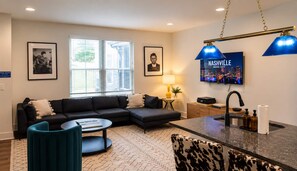 A modern living room with a sectional sofa, a round coffee table, a TV mounted on the wall, and a kitchen area with a sink and countertop. - Sleeps 8 near Broadway — Hart Suites Unit #6 (Nashville)