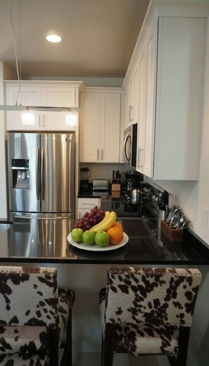 A modern kitchen with stainless steel appliances, a fruit bowl on the counter, and two patterned chairs. - Sleeps 8 near Broadway — Hart Suites Unit #6 (Nashville)