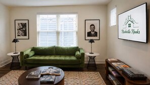 Een woonkamer met een groene bank, twee bijzettafels, een salontafel met boeken, ingelijste foto's en een bordje aan de muur.