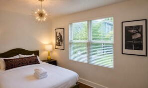 A hotel room with a bed, a nightstand, a lamp, a window with blinds, and two framed pictures on the wall. - Free parking near Broadway — Hart Suites #7 (Nashville)