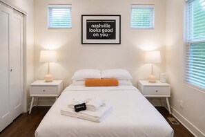 Ein Hotelzimmer mit einem ordentlich bezogenen Bett, zwei Nachttischen mit Lampen, einem gerahmten Zitat an der Wand und einem Fenster mit Jalousien.