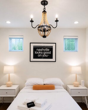 A hotel room with a bed, two bedside tables, a chandelier, two windows with blinds, and a framed quote on the wall. - Sleeps 8 near Broadway — Hart Suites #14 (Nashville)