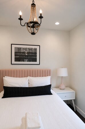 A hotel room with a bed, a nightstand, a lamp, a framed picture on the wall, and a chandelier. - 10 Min to Broadway | Sleeps 8 | Hart Suites #16 (Nashville)