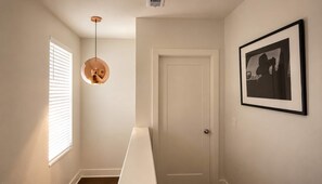 Een gang met een witte deur, een ingelijst schilderij aan de muur en een hanglamp.