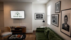 A living room with a flat-screen TV, a green sofa, a wicker chair, a coffee table with magazines, and framed pictures on the wall. - Spacious townhome, sleeps 8 — Hart Suites #8 (Nashville)