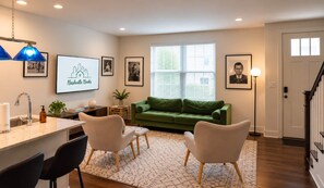 A living room with a green sofa, two white chairs, a coffee table, a TV displaying a cityscape, framed pictures on the wall, a window with blinds, and a door. - Spacious townhome, sleeps 8 — Hart Suites #8 (Nashville)