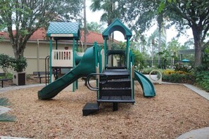 Une aire de jeux avec un toboggan et une structure d’escalade, entourée d’arbres et bordée d’un bâtiment dont le toit est recouvert de tuiles rouges.