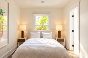 A bedroom with a large bed, two bedside tables with lamps, a window with a view of greenery, and a door to the right. - Luxury 1913 Craftsman in Venice Beach (Los Angeles)