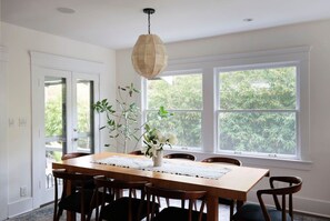 A dining area with a wooden table, chairs, a hanging light fixture, and large windows with a view of greenery. - Luxury 1913 Craftsman in Venice Beach (Los Angeles)