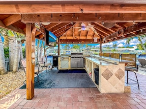 An outdoor kitchen area with a grill, bar counter, and seating under a wooden structure. - Pool & Hot Tub! Waterfront+Close To Beach! (Pompano Beach)