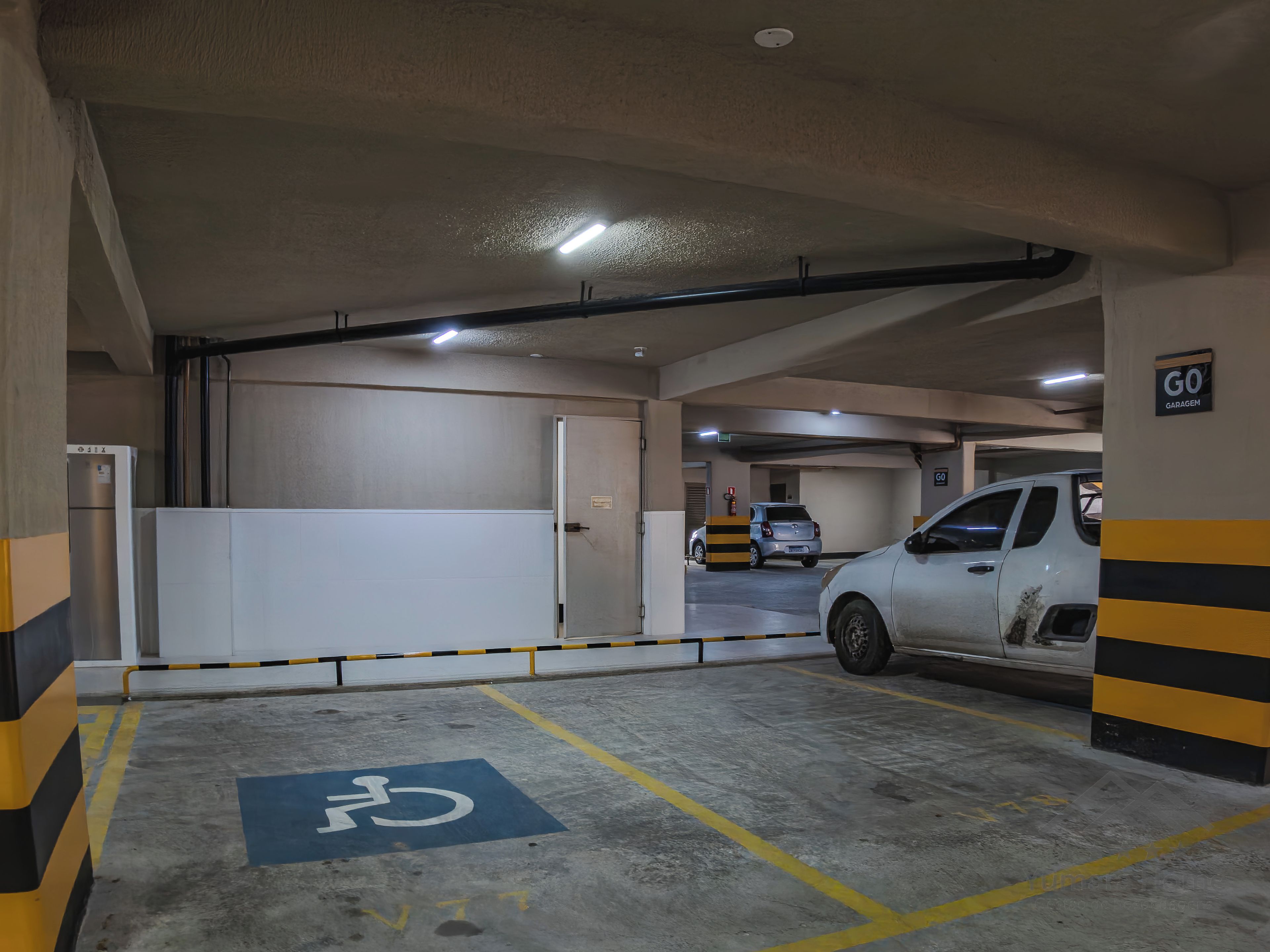 Een parkeergarage met een gehandicaptenparkeerplaats, een witte auto op de parkeerplaats en een 'GO GARAGEM'-bord aan de muur.