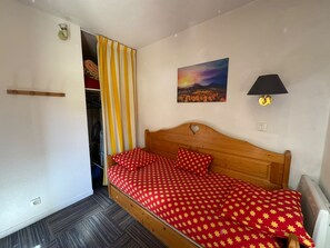 A bedroom with a wooden bed, a red and yellow patterned bedspread, a painting of a sunset, a black lamp, and a closet with yellow and white striped curtains. - Séjour montagne - 6 pers avec balcon à Risoul (Risoul)