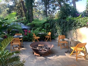 Ein Sitzbereich im Freien mit Feuerstelle, umgeben von Stühlen und Tischen, eingebettet in einen üppigen Garten mit Bäumen und Pflanzen.