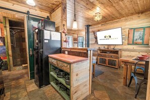Une cuisine rustique avec un îlot central en bois, un réfrigérateur noir et un téléviseur diffusant l’émission « ModernVacay ».