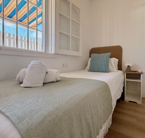 Een netjes opgemaakt bed met een groene deken, witte kussens en een blauw kussen. Er staat een klein nachtkastje met een lamp en een stopcontact.