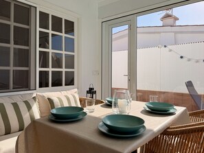 Un coin repas avec une table dressée pour deux personnes, une chaise en osier et une vue sur une clôture blanche et un bâtiment en arrière-plan.
