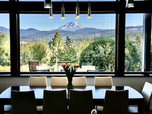 A dining area with a table and chairs, a vase with flowers, and a view of a mountain through large windows. - Phato's Retreat - Luxury Home, Hot Tub, Stunning Views (White Salmon)