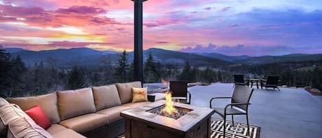 An outdoor seating area with a fire pit, a sofa, and chairs, overlooking a mountainous landscape.