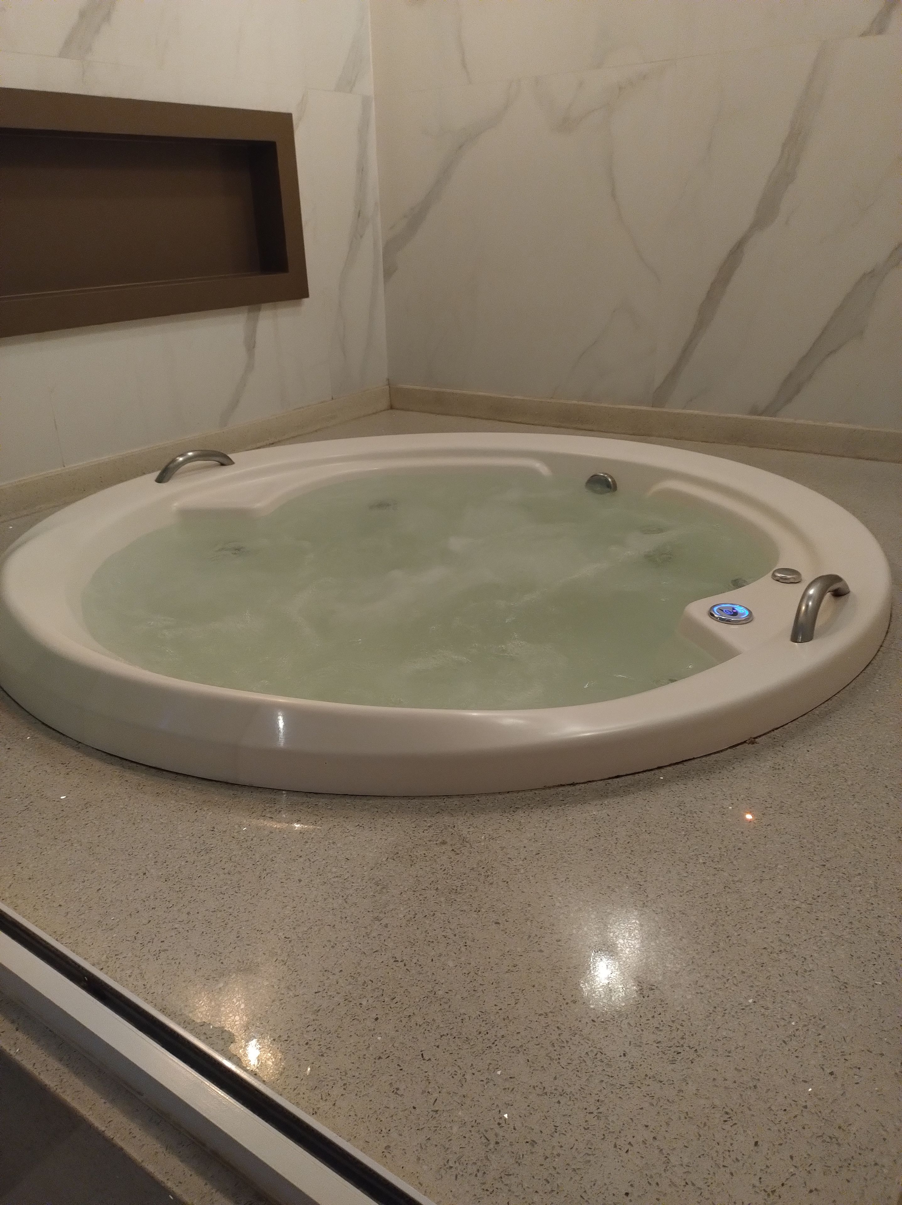A round bathtub with a control panel, set on a speckled floor, against a wall with a marble pattern.