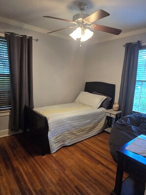 Un dormitorio con cama, mesita de noche, ventilador de techo y ventana con cortinas.