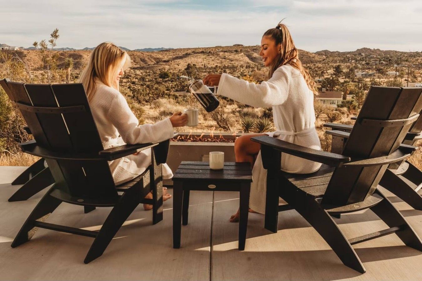 Deux personnes sont assises à une table entourée de chaises, et elles ont devant elles un paysage vallonné avec une végétation clairsemée.