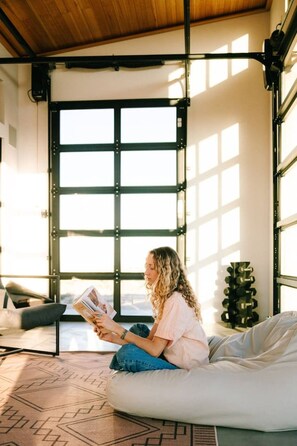 Une personne est assise sur un fauteuil en forme de sac de haricots, en train de lire un magazine dans une pièce dotée de grandes fenêtres et dont on aperçoit la porte.