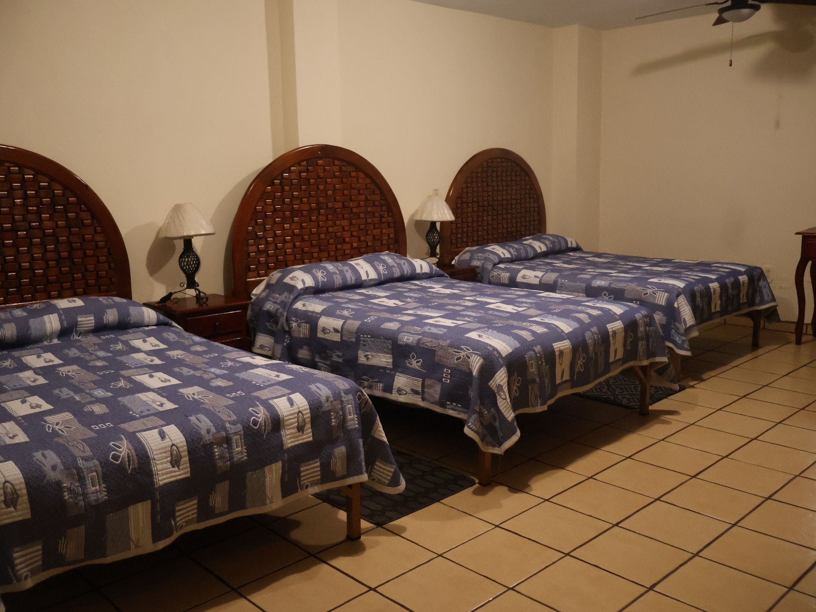 A hotel room with three wooden beds, each with a patterned bedspread, a nightstand with a lamp, and a tiled floor.