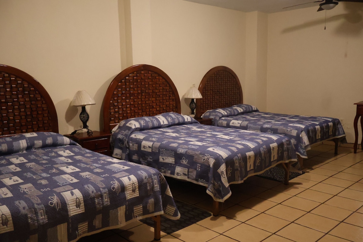 A hotel room with three wooden beds, each with a patterned bedspread, a nightstand with a lamp, and a tiled floor.