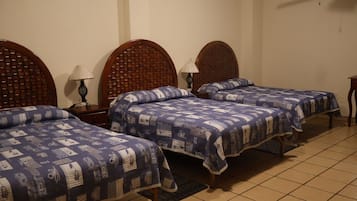 A hotel room with three wooden beds, each with a patterned bedspread, a nightstand with a lamp, and a tiled floor.