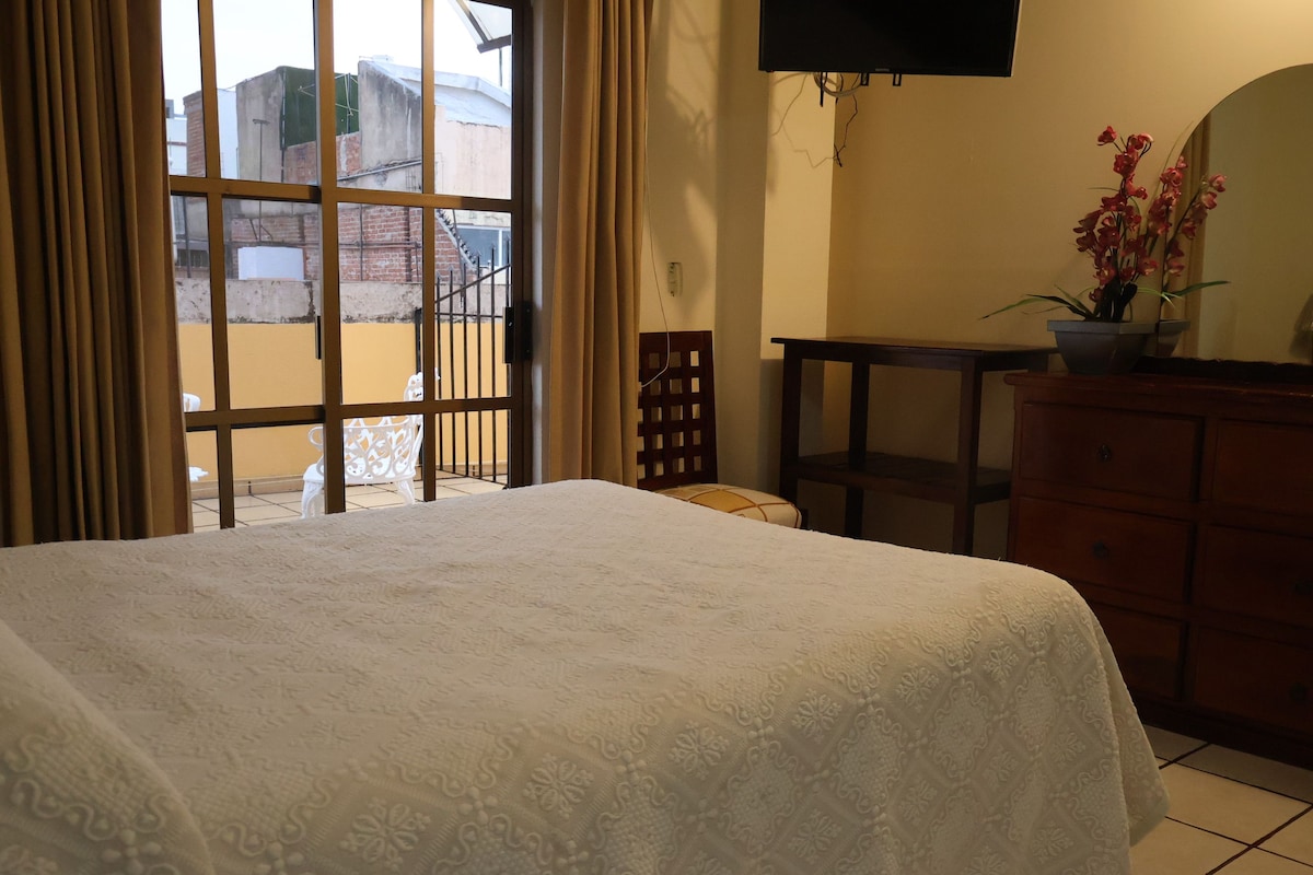 A hotel room with a bed, a TV mounted on the wall, a small table, a dresser, and a view through a window.