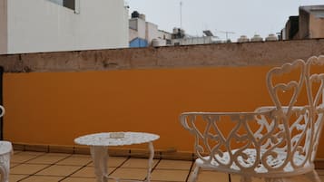 Una terraza en la azotea con una mesa y una silla de hierro forjado blanco, suelo de baldosas y una pared naranja lisa.