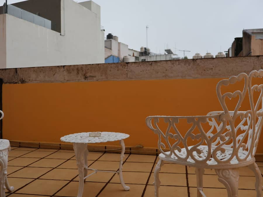 Una terraza en la azotea con una mesa y una silla de hierro forjado blanco, suelo de baldosas y una pared naranja lisa.
