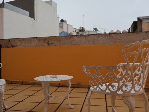Una terraza en la azotea con una mesa y una silla de hierro forjado blanco, suelo de baldosas y una pared naranja lisa.