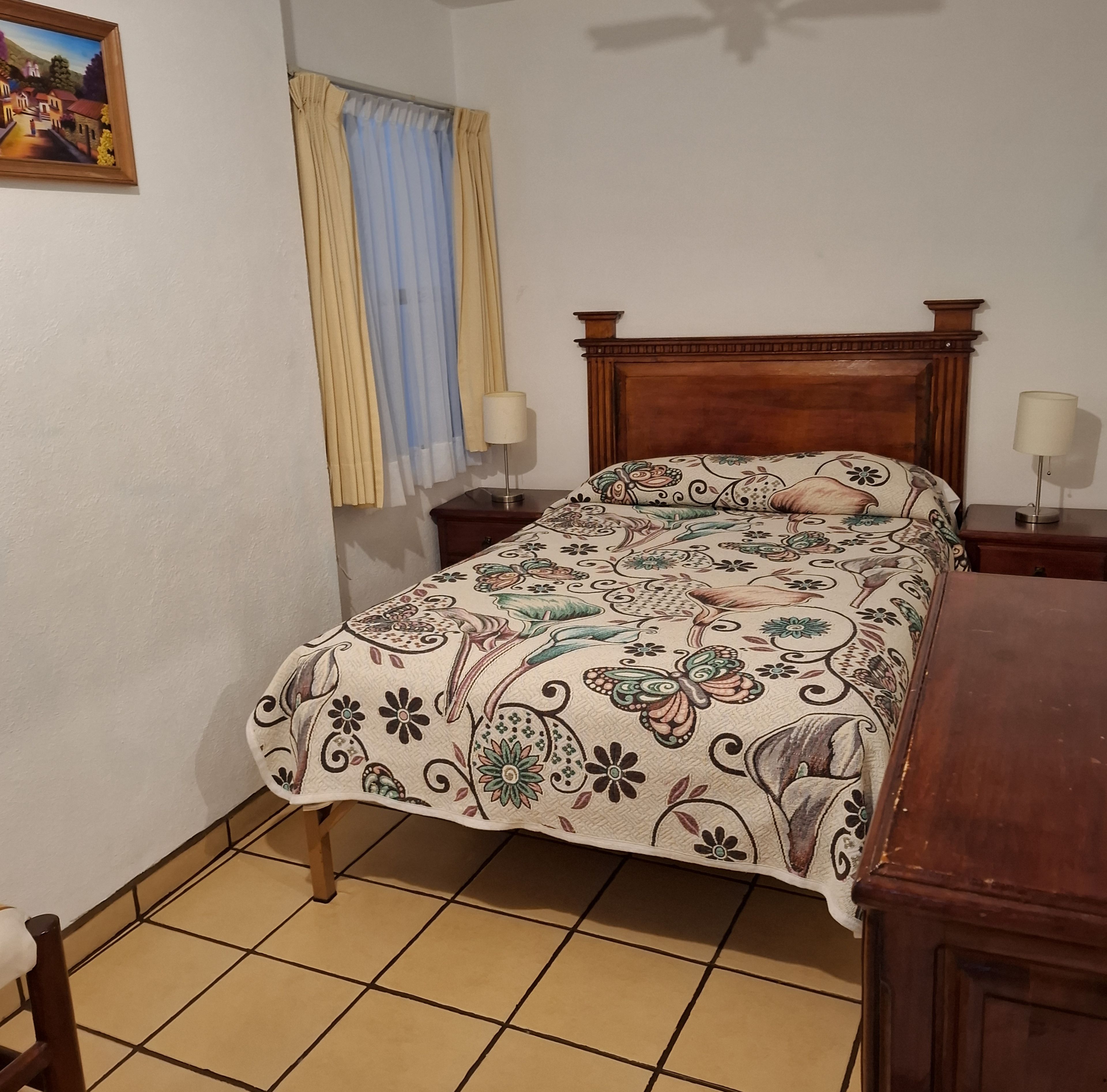 Une chambre à coucher avec un lit, deux tables de chevet, une commode, une fenêtre avec des rideaux et un tableau au mur.