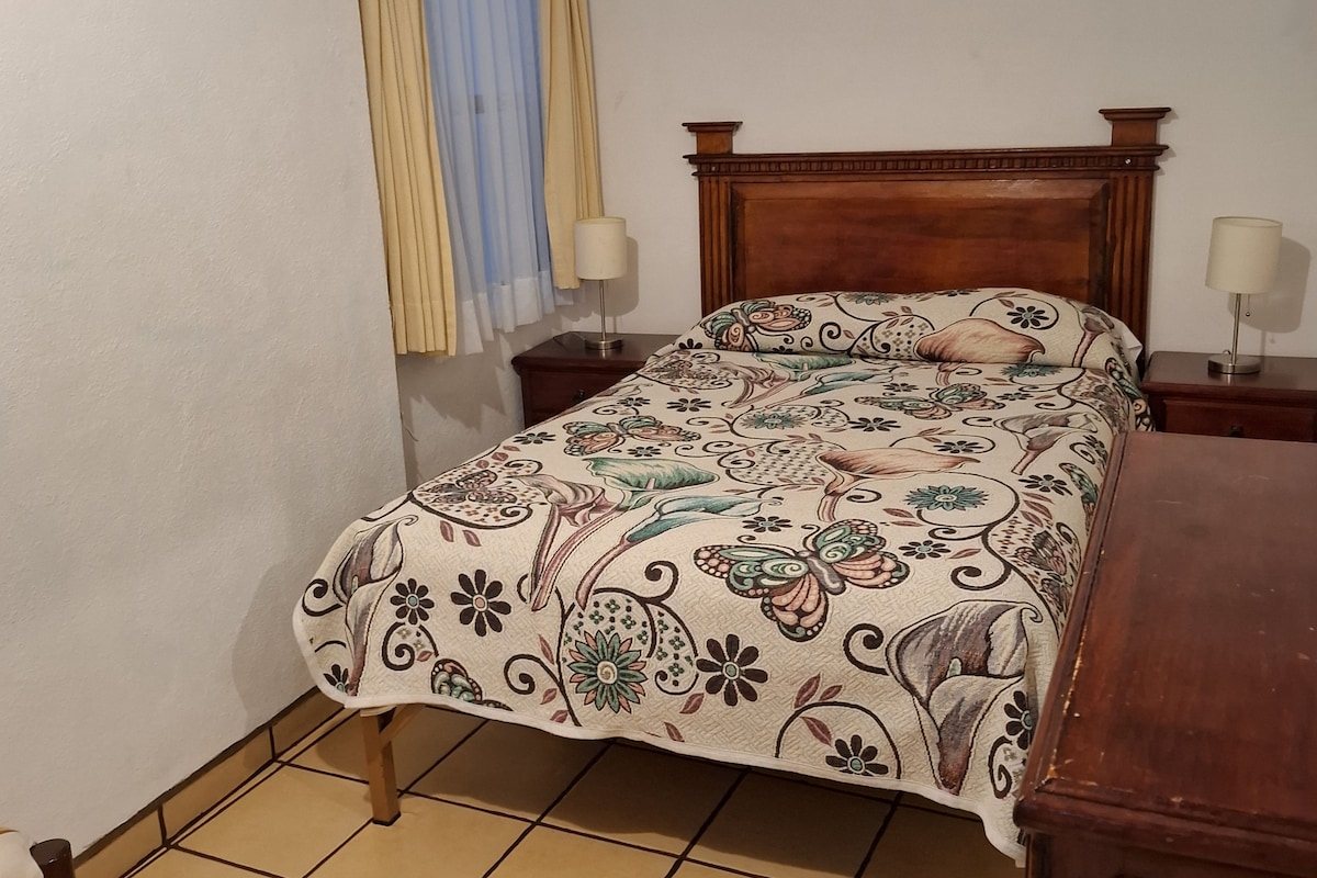 A bedroom with a bed, two bedside tables, a nightstand, a window with curtains, and a painting on the wall.