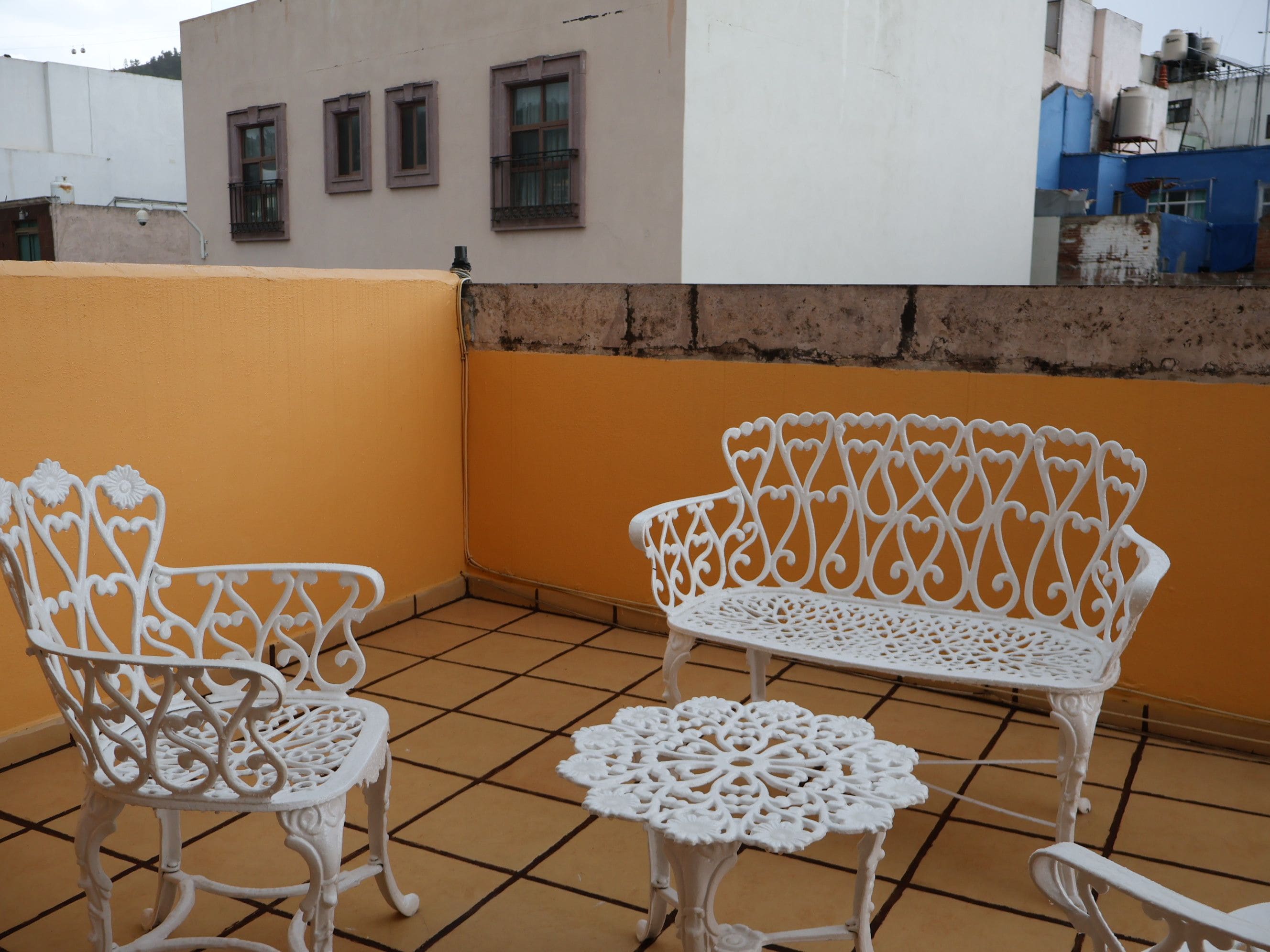 A balcony with two ornate white metal chairs and a small round table, with a tiled floor and a building in the background.