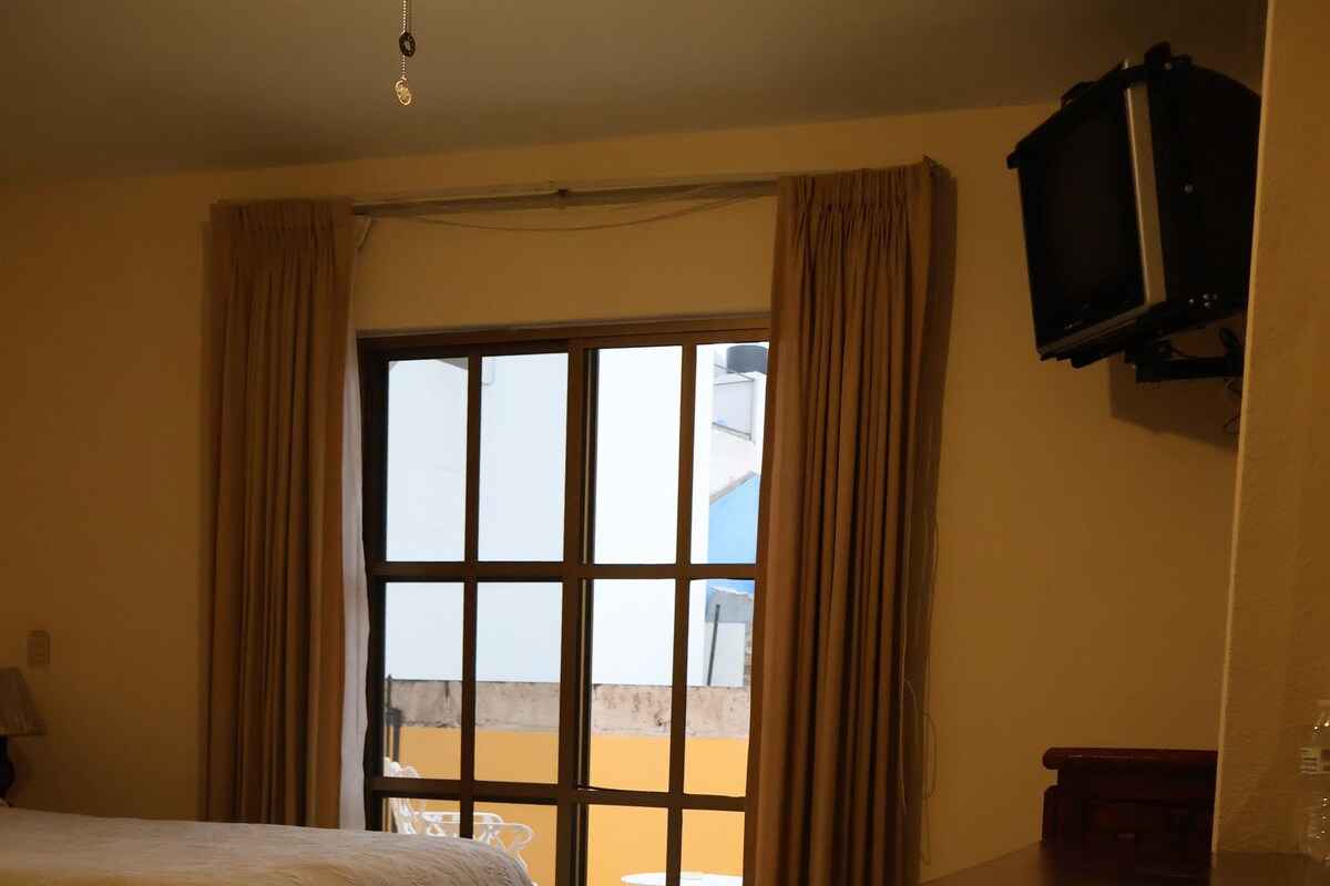A hotel room with a bed, a desk, a television mounted on the wall, and a window with curtains.