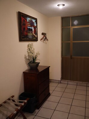 Una habitación con piso de baldosas, cómoda de madera, cuadro enmarco, planta en maceta, perchero colgante y puerta corrediza.