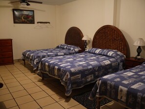 Un dormitorio con tres camas, un aparador de madera, un ventilador de techo, un cuadro colgado en la pared y una alfombra estampada.