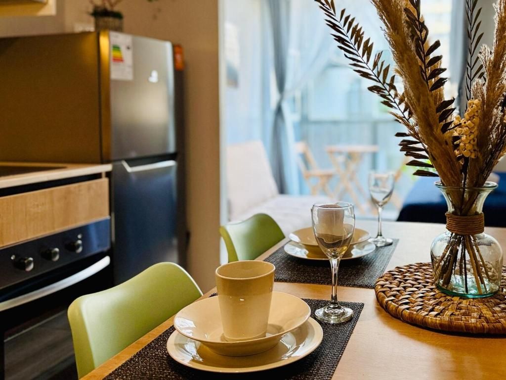 Un comedor con una mesa preparada para dos, un jarrón con plantas secas y una cocina al fondo.