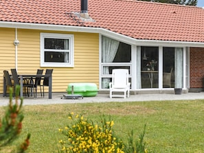 Una casa amarilla con techo de tejas rojas, un patio con mesa y sillas, y un bote de basura verde.