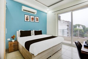 A bedroom with a bed, a nightstand, a vase with flowers, and a window with a view of buildings. - Super Townhouse Subhash Chowk (Gurugram)